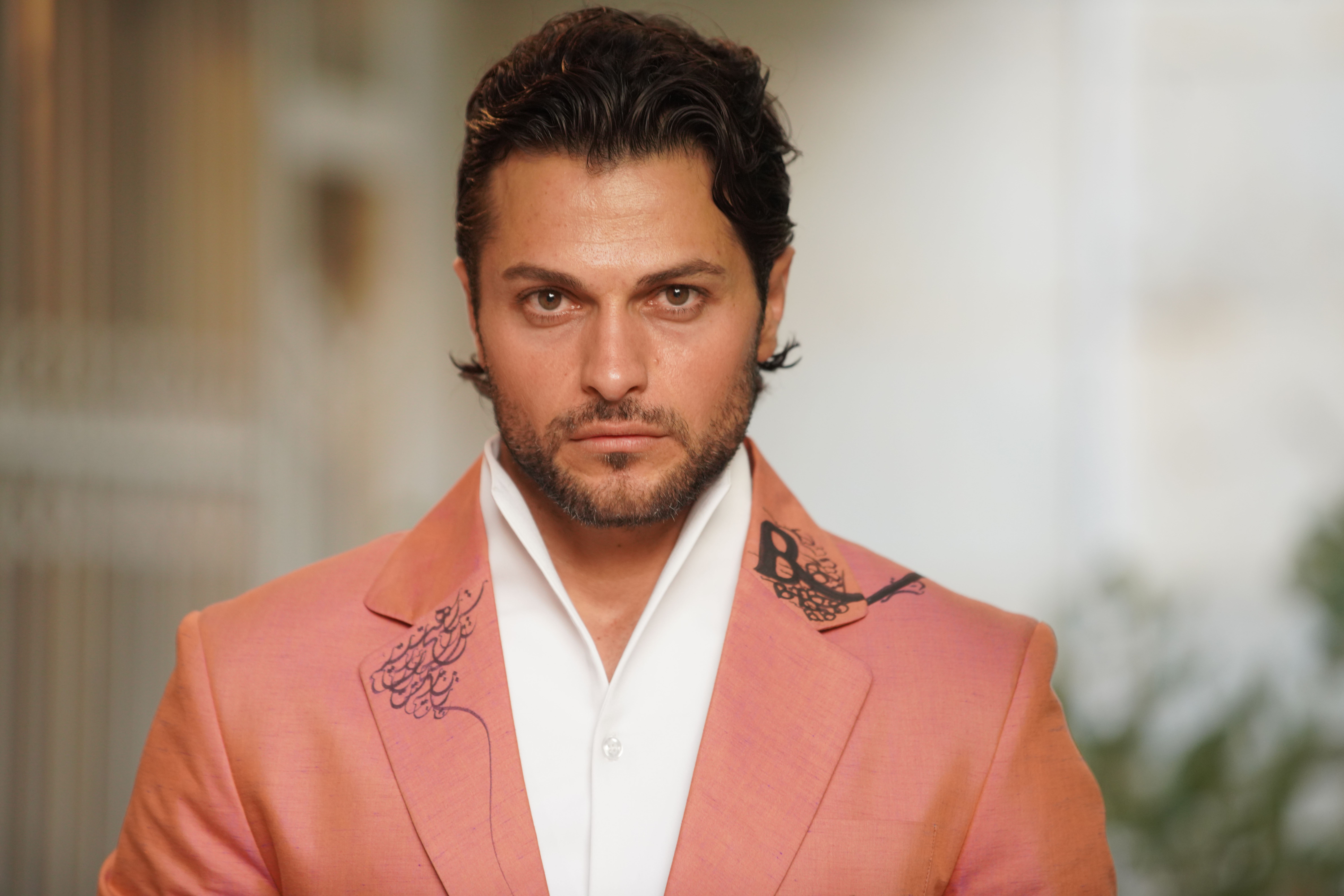 A confident man poses in a stylish pink suit against a blurred outdoor backdrop.