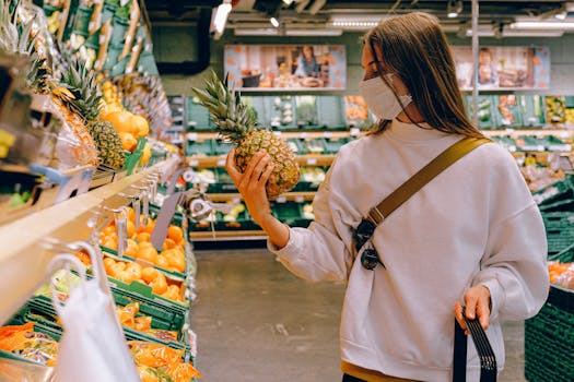 Woman in mask chooses pineapple in a supermarket for safe shopping during pandemic.