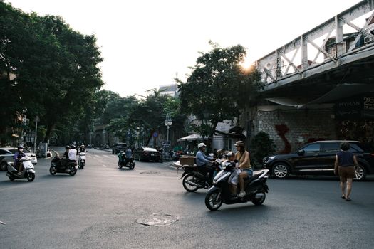 Motorbikes navigate a lively urban street under a railway bridge at sunset.