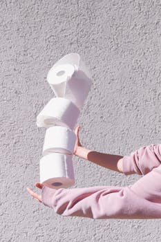 A hand balances a stack of toilet rolls against a textured wall in bright lighting.
