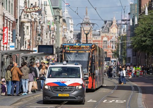Free Lively Amsterdam street scene featuring tram, police van, and people at a tram stop. Stock Photo