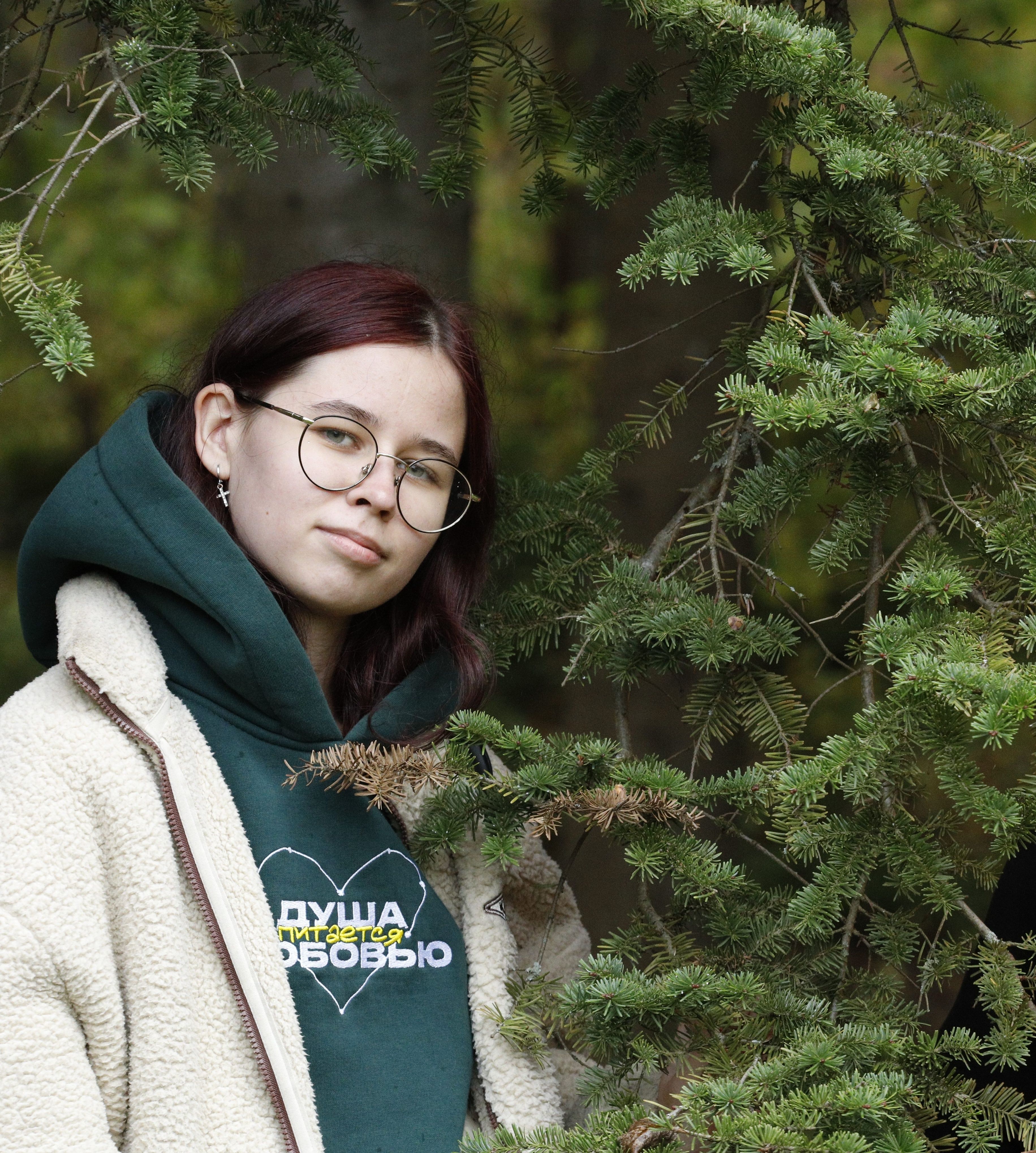 A young woman in glasses and a hoodie smiles gently among pine trees outdoors.