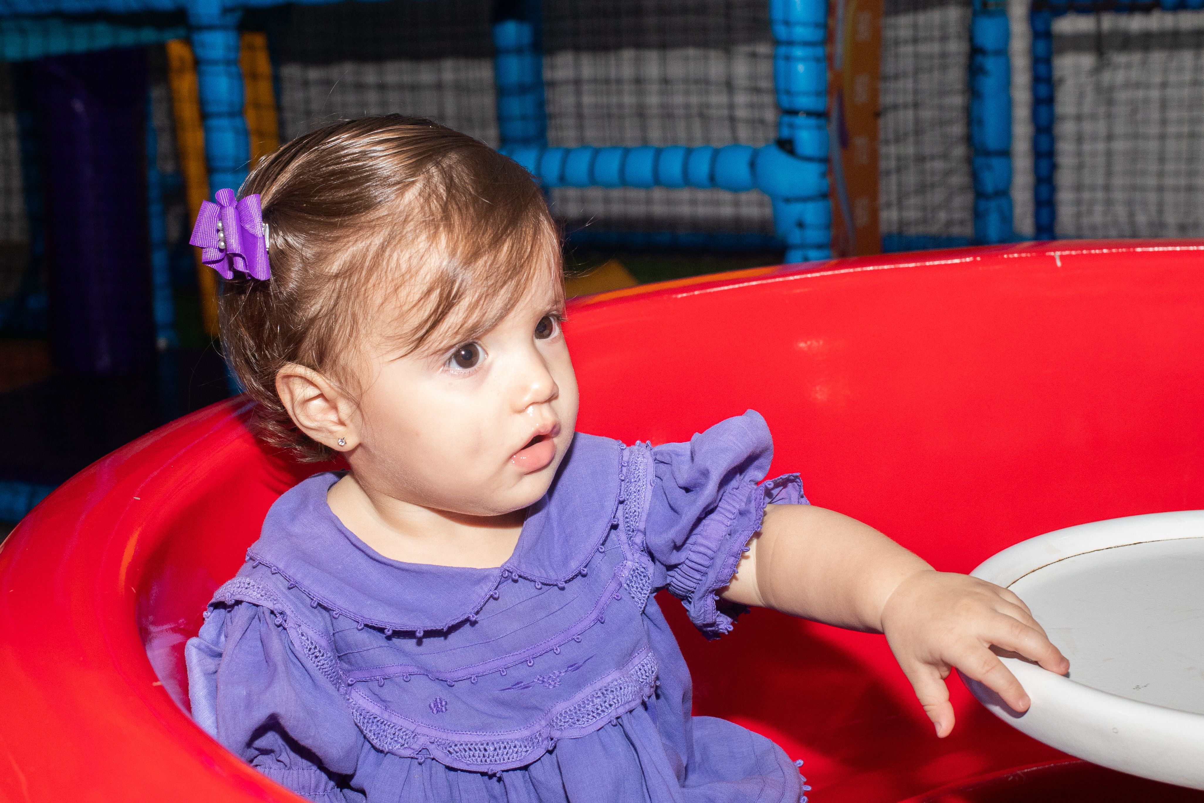 A cute baby wearing a purple dress plays in a colorful indoor play area, exploring a red ball pit.