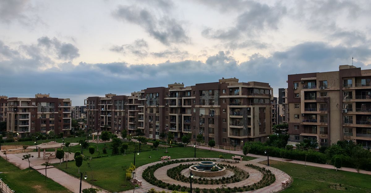 Aerial view of modern apartment buildings surrounding a landscaped garden with a fountain at dusk.