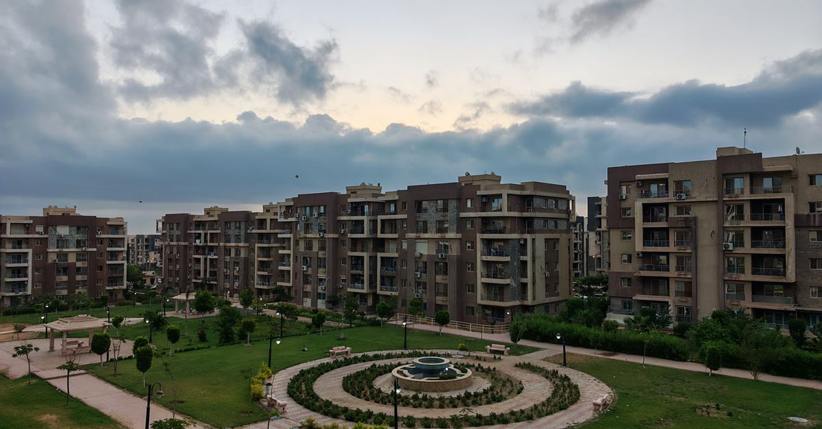 Modern apartment buildings surround a lush central park with a fountain under a cloudy sky.