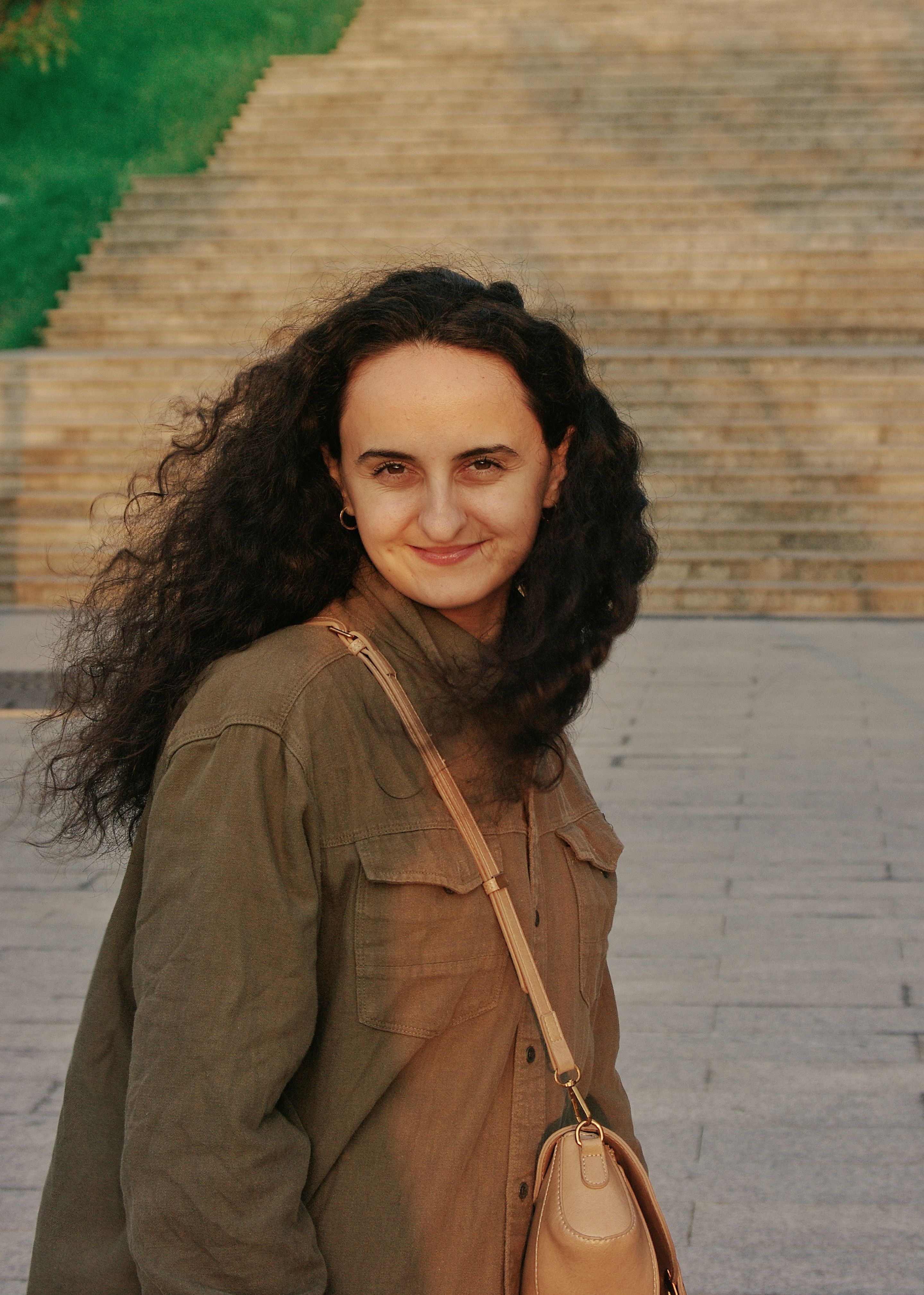 A young woman with curly hair smiles while standing on urban steps in daylight.