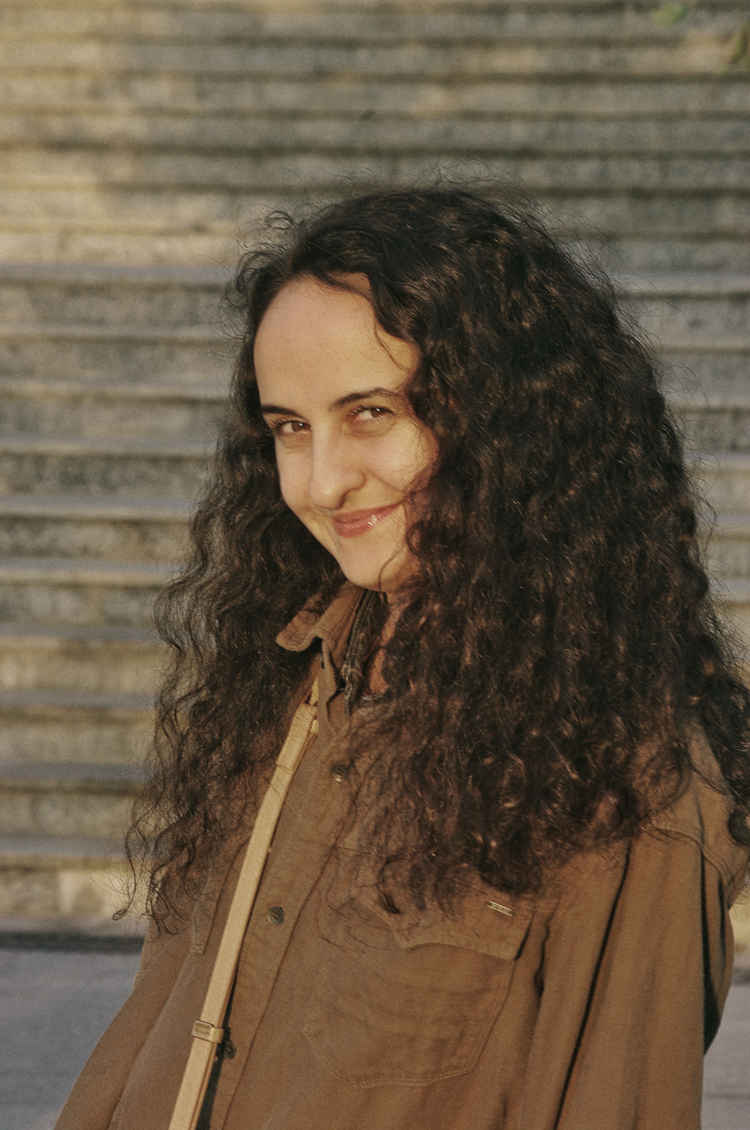 A woman with curly hair smiles warmly on sunny outdoor steps.