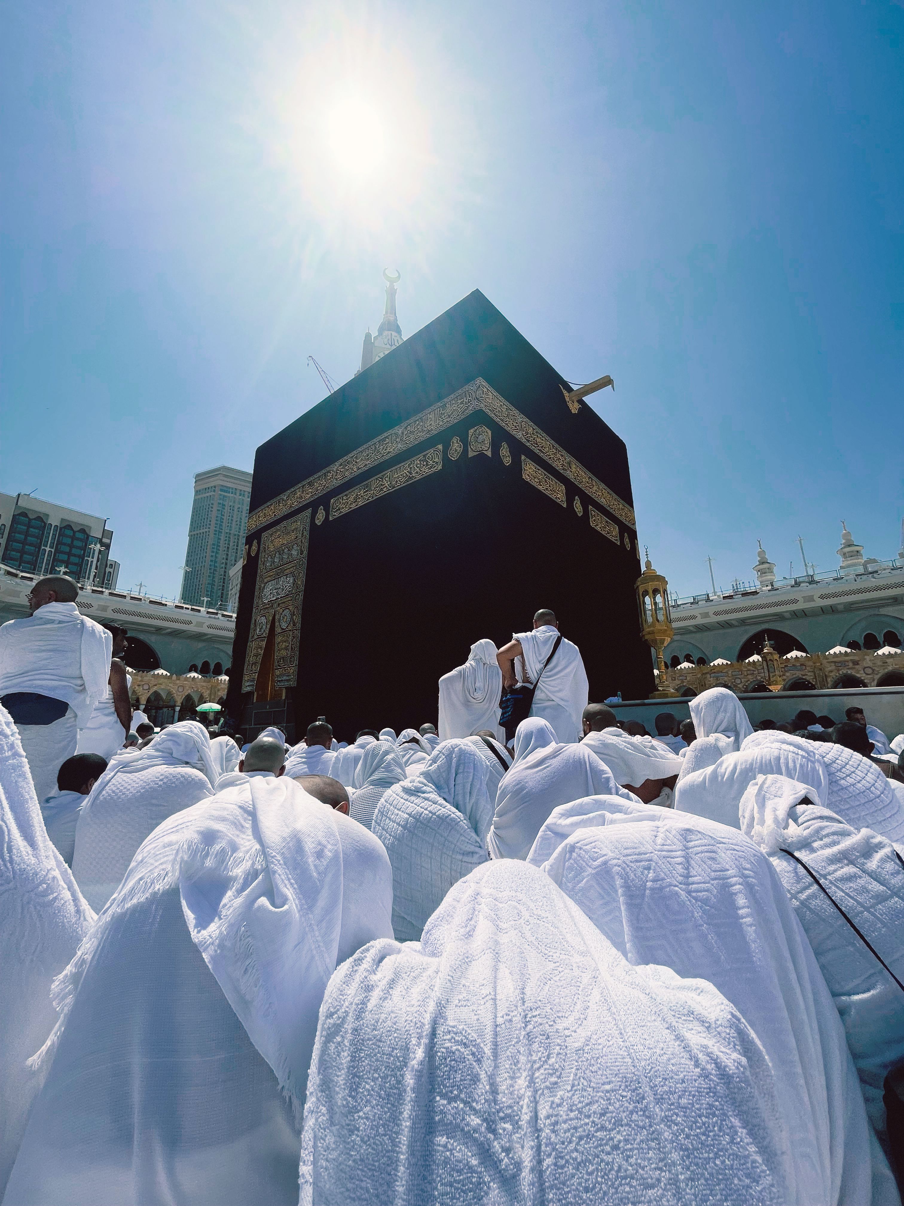 Group of pilgrims praying around the Kaaba in Makkah under a bright sun.