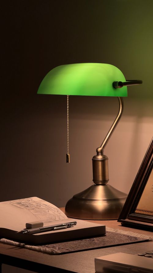 Free A vintage banker's lamp with a green shade and brass base illuminates a cozy desk scene. Stock Photo