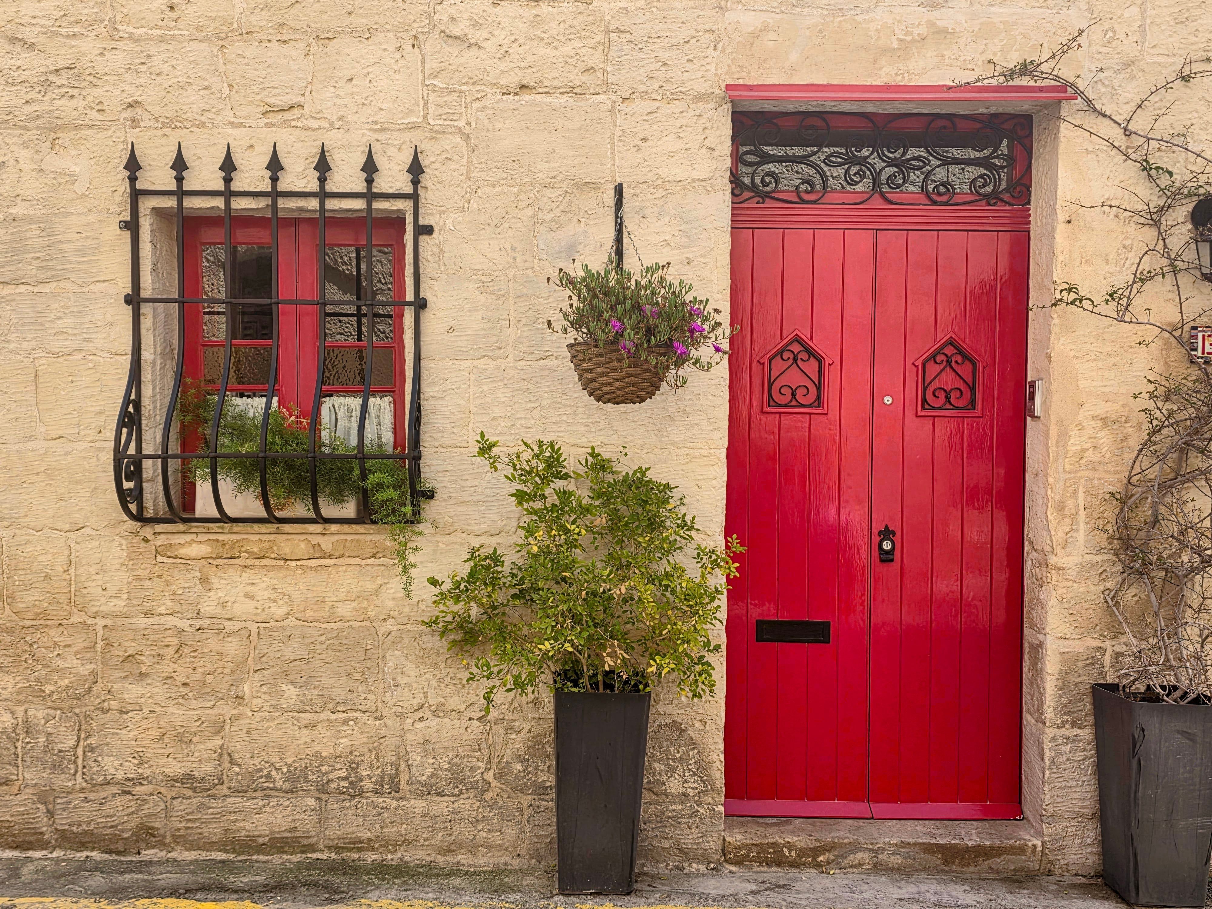 A striking red door and window on a stone building in Rabat, Malta, with plants.