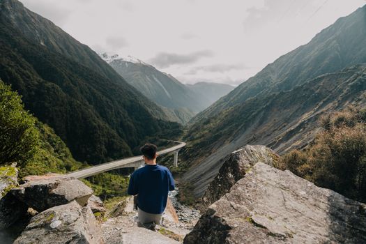 A man stands on rocky terrain, overlooking a scenic mountain pass and winding road.