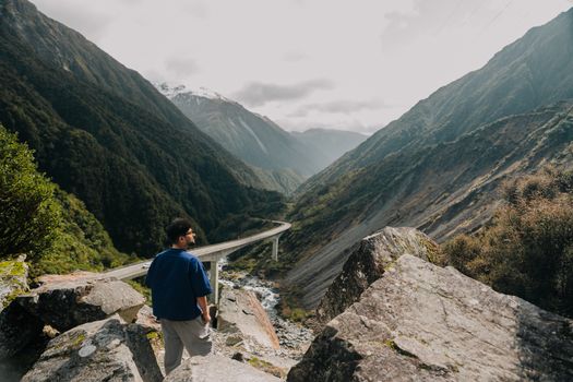 A man stands on rocks, admiring a stunning mountain valley view with a distant bridge.