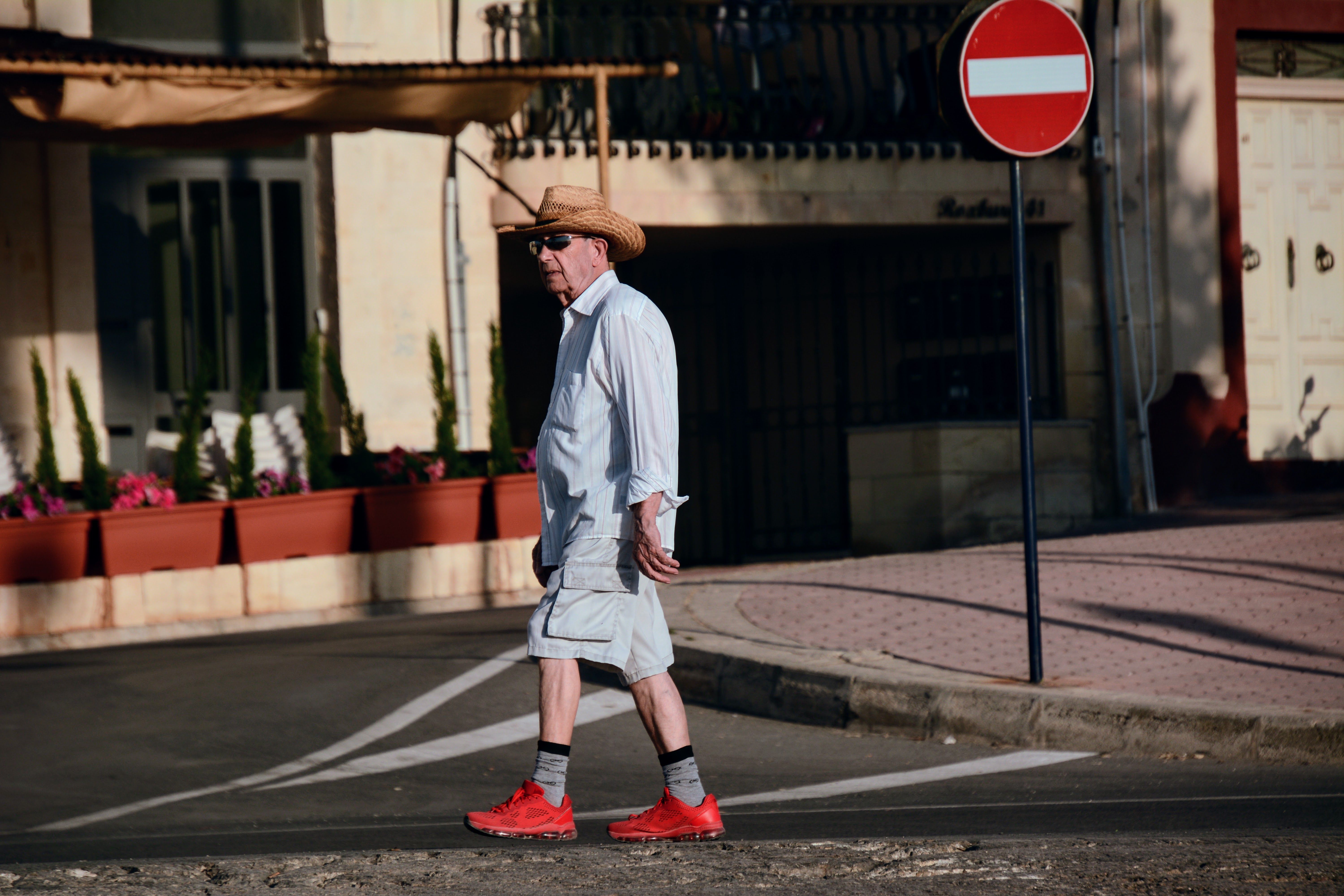 Elderly man in a straw hat and red shoes crossing a sunny street with a do not enter sign.