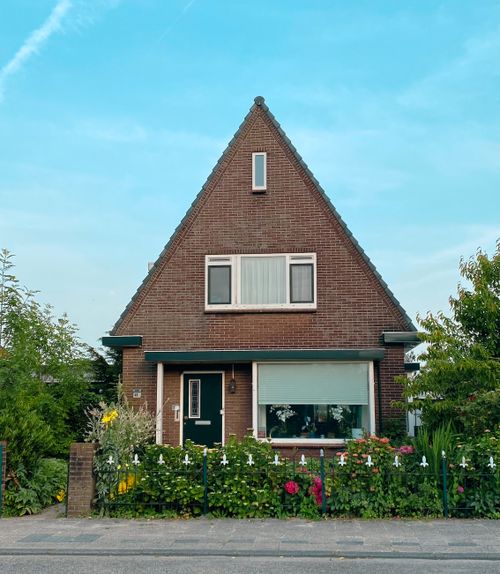 Free A picturesque Dutch brick house with a triangular roof, surrounded by a vibrant garden under a clear blue sky. Stock Photo
