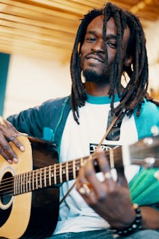 A portrait of an African American man with dreadlocks playing an acoustic guitar indoors.