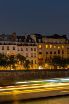 Elegant Parisian buildings by the river at night with light trails from a passing boat, showcasing urban beauty.