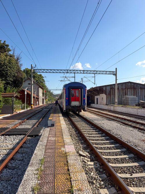 Free A solitary train exits a quiet rural station under a clear blue sky, surrounded by greenery. Stock Photo