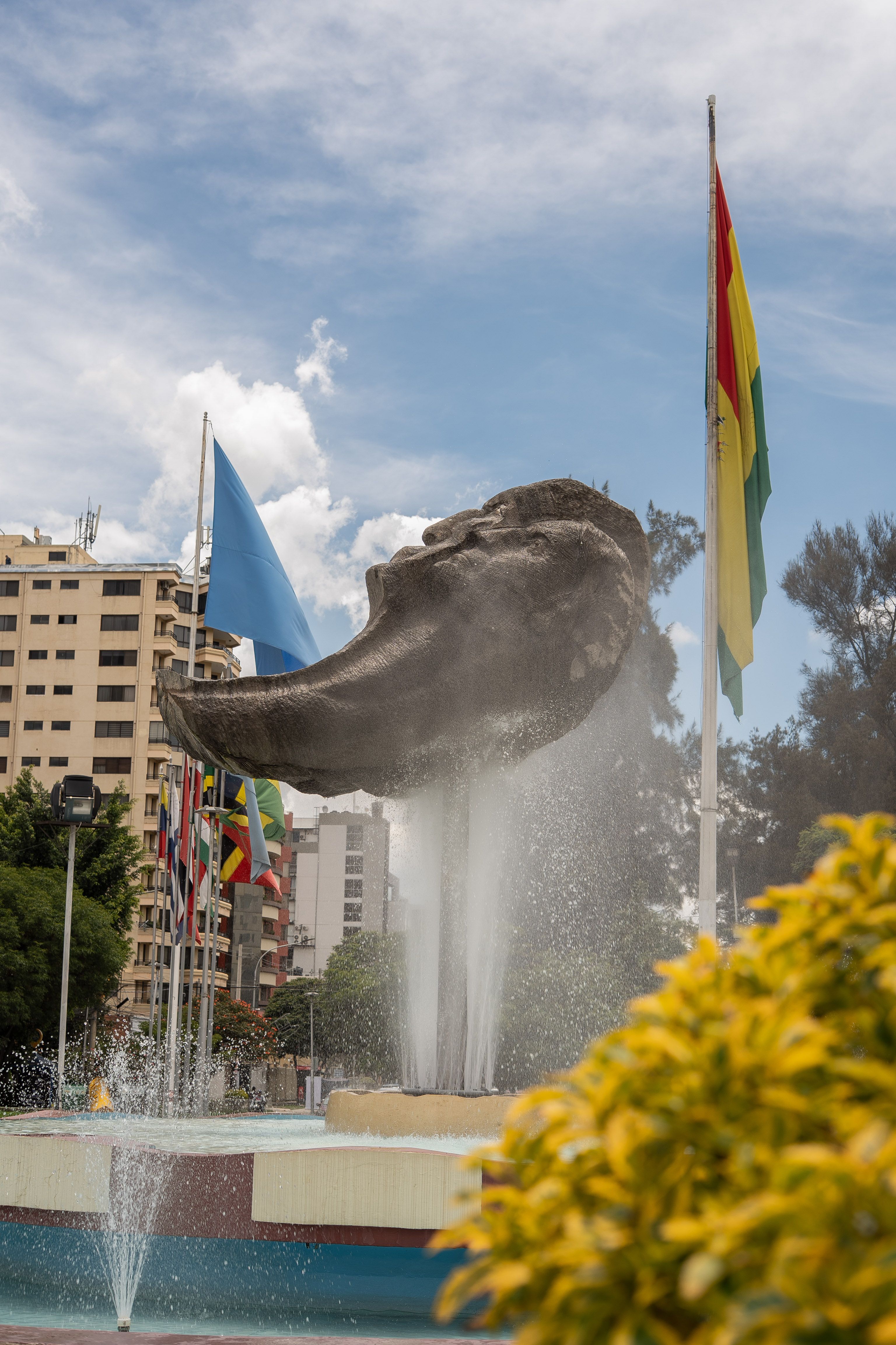 Sculpture surrounded by flags in a city plaza, clear sky above.