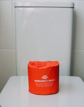 Minimalist shot of an emergency toilet paper roll in vivid red packaging placed on a toilet lid indoors.