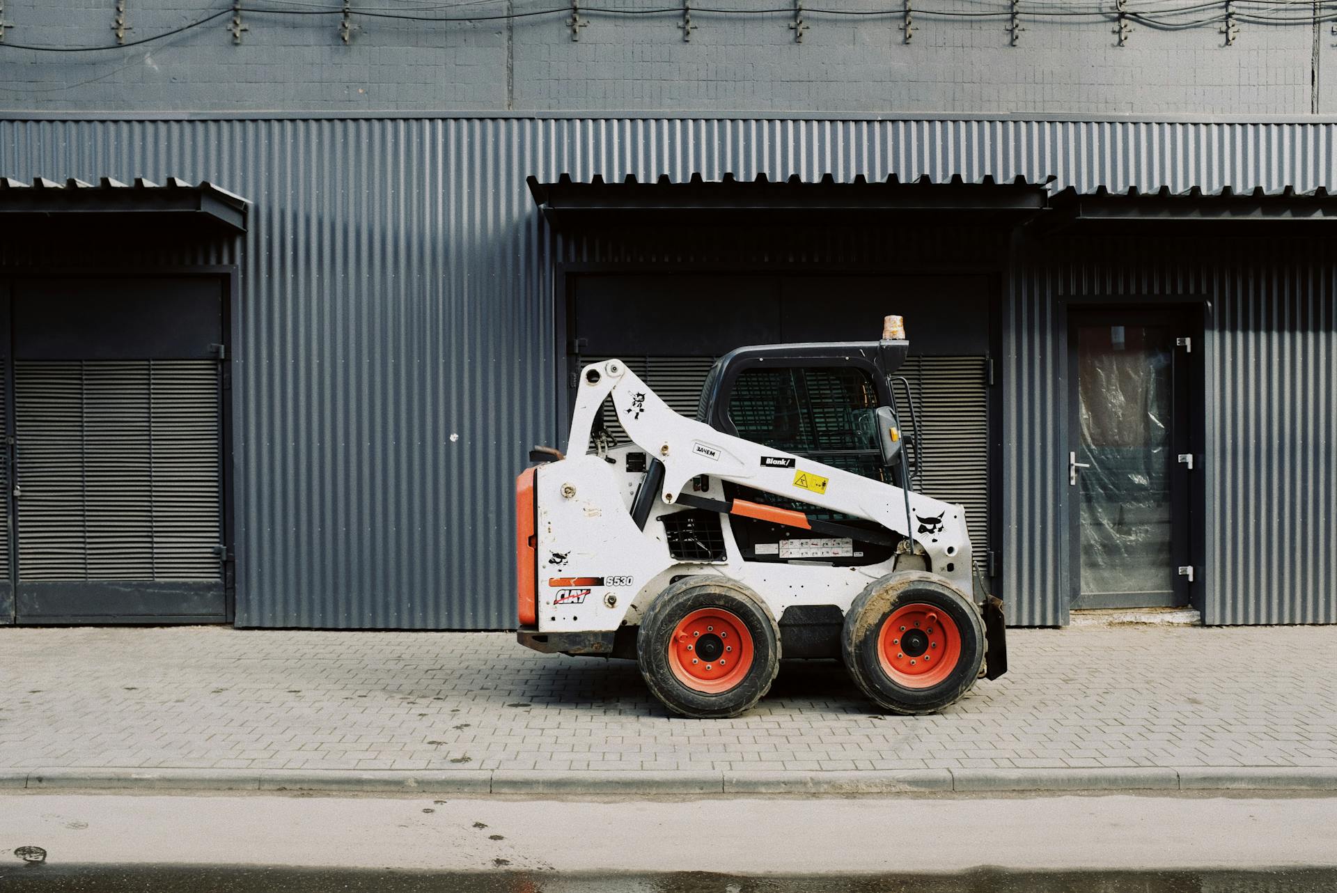 White skid steer loader parked outside a corrugated metal warehouse in an urban area during the day.