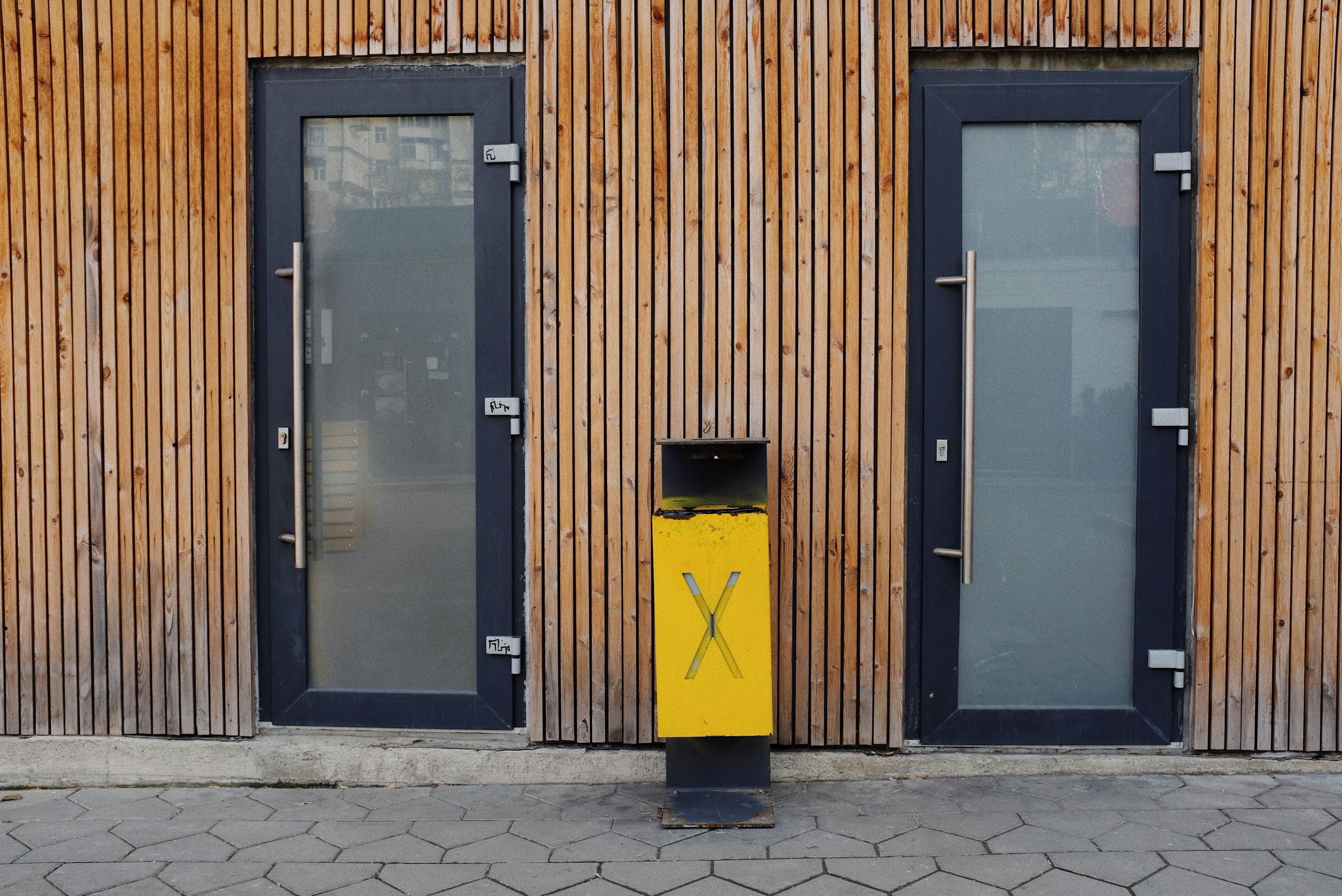 A modern exterior featuring wooden panels, glass doors, and a yellow trash bin in an urban setting.