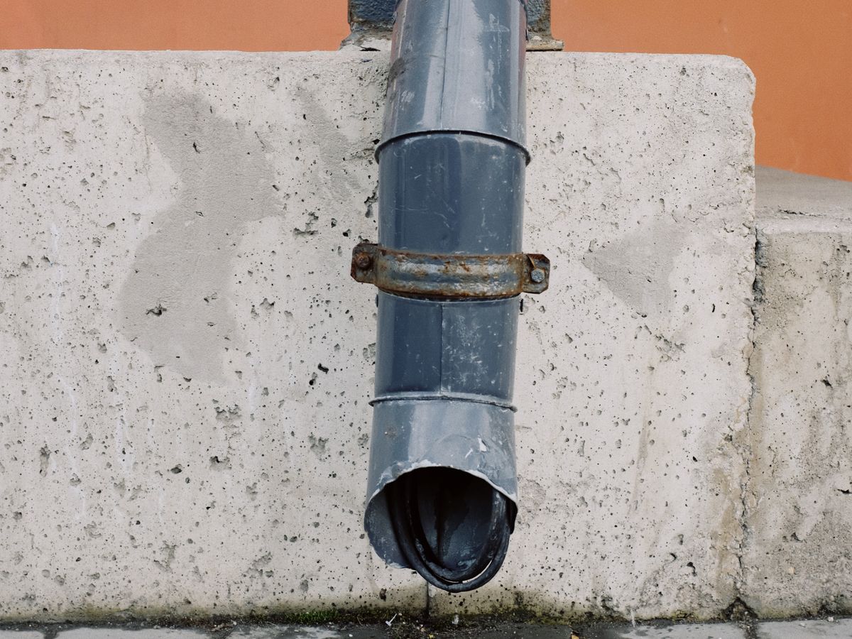 A close-up of a metal house drain pipe on a concrete wall with water runoff