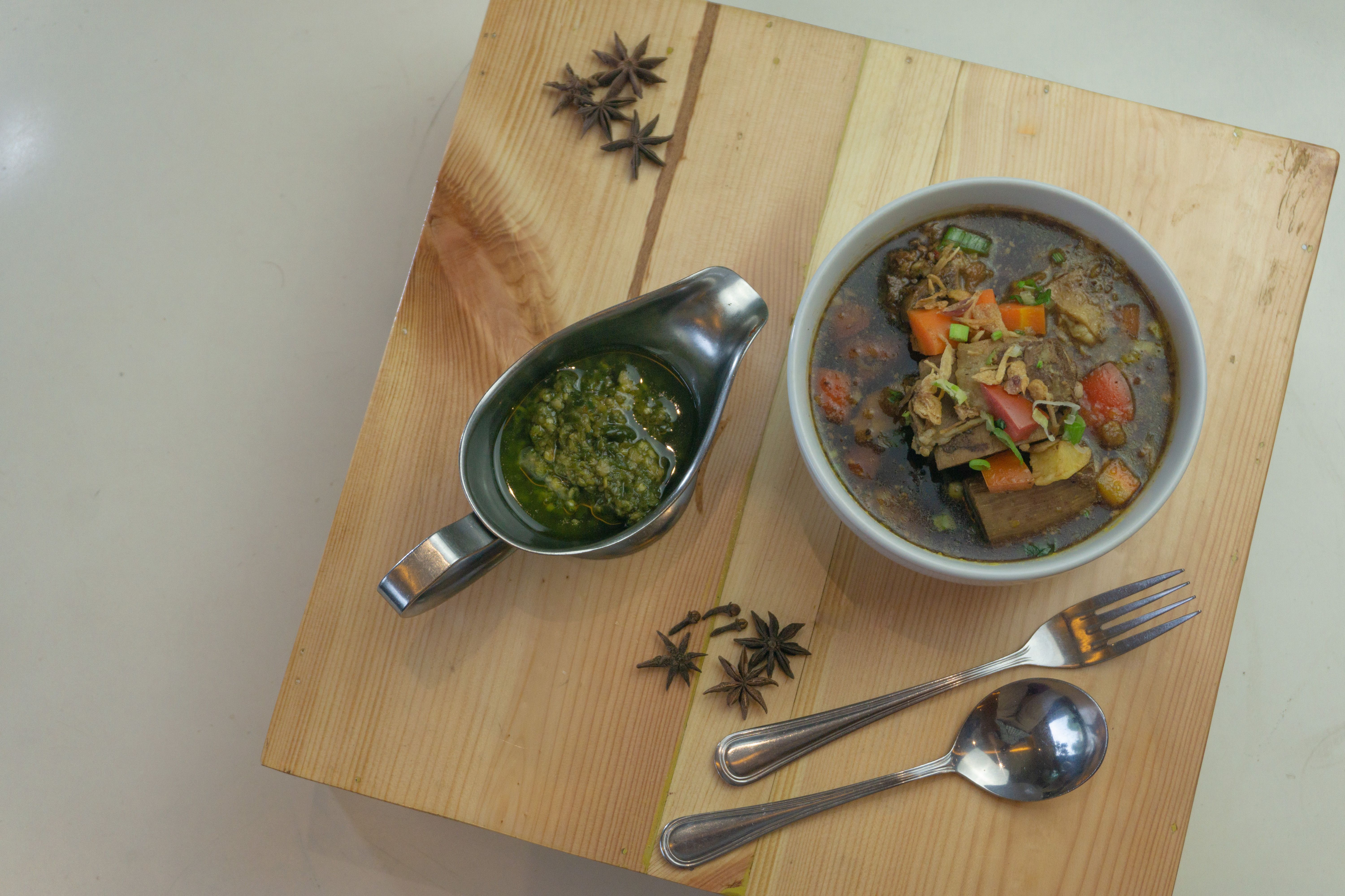 Flat lay of a flavorful Asian soup with herb sauce garnished with star anise, and cutlery on a wooden board.