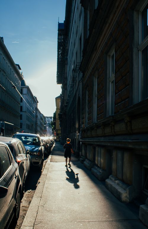 Free A person walks along a sunlit city street lined with cars and historic buildings. Stock Photo