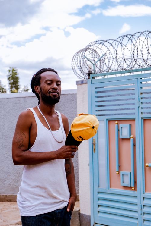 Free Man in casual outfit standing by a colorful gate with barbed wire, holding a cap. Representation of urban lifestyle. Stock Photo