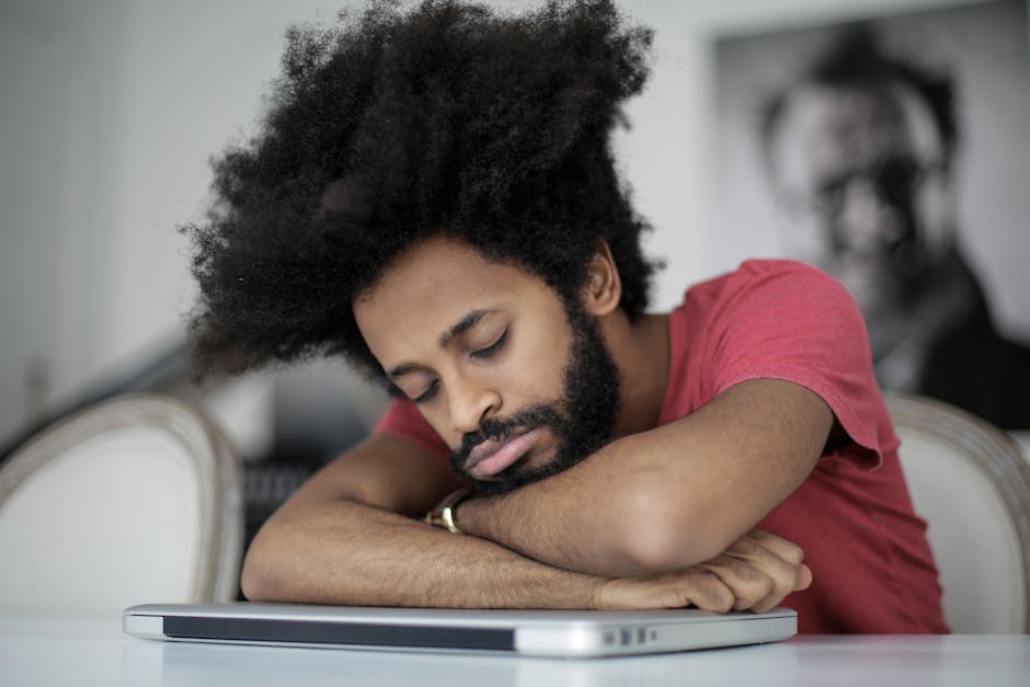 Foam vs Silicone vs Custom Earplugs: Which Best Blocks Noise for Sleep? Man with afro hair sleeping on laptop, symbolizing remote work fatigue.