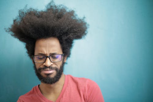 Young bearded ethnic male with creative Afro hairstyle wearing eyeglasses and pink t shirt looking down pensively thinking about trouble or question