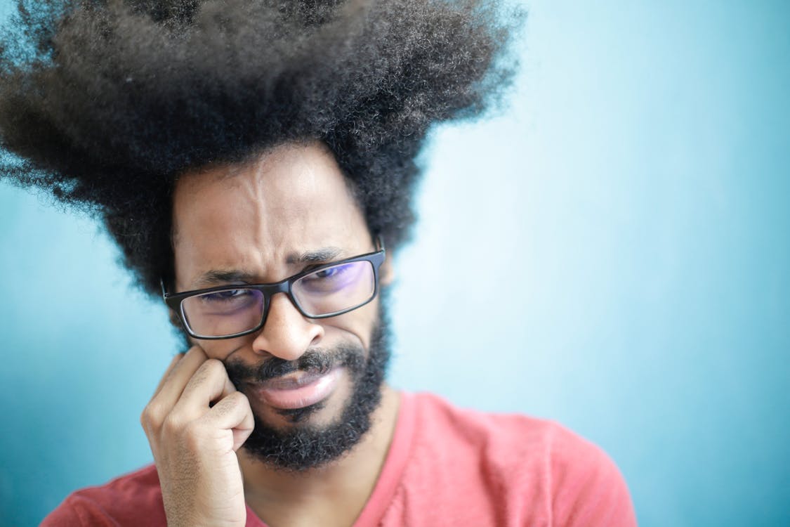 Suspicious young bearded ethnic male with creative Afro hairstyle wearing eyeglasses and pink t shirt touching cheek and looking at camera with uncertain or doubt expression