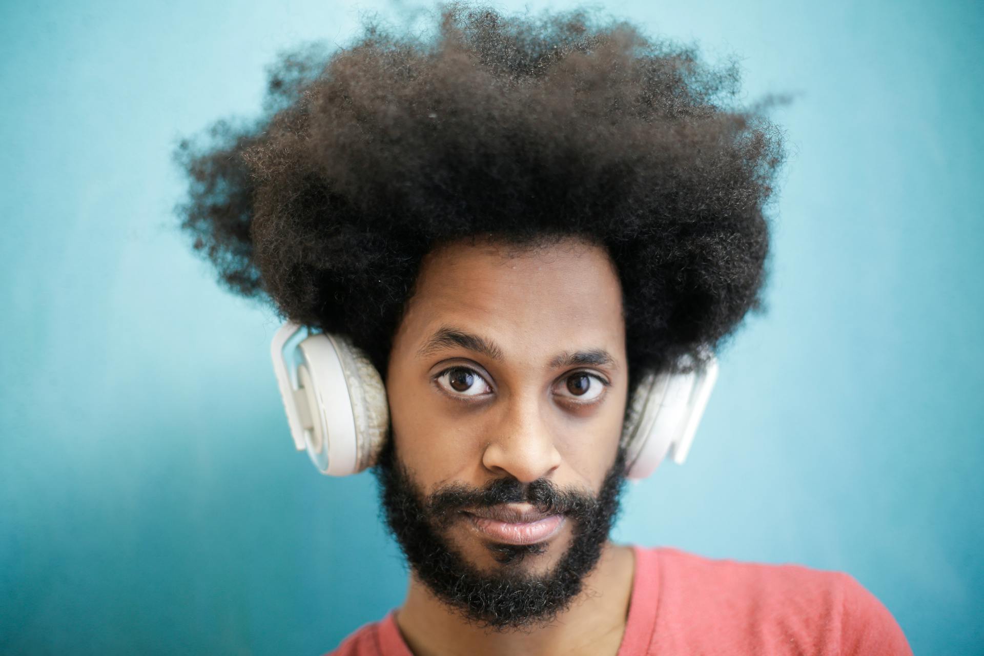 Calm positive young ethnic male with creative afro hairstyle wearing pink t shirt using white headphones and looking at camera