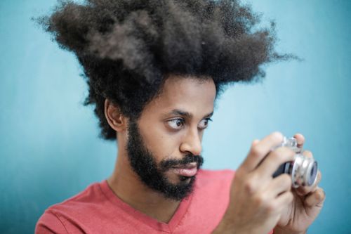 Free Ethnic male with creative Afro hairstyle wearing pink t shirt taking pictures on vintage photo camera against blue background while looking away thoughtfully Stock Photo