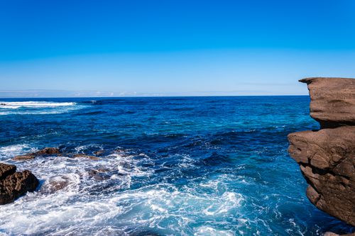 Free Vivid blue ocean waves crash against rocky coastal cliffs under a clear sky. Stock Photo