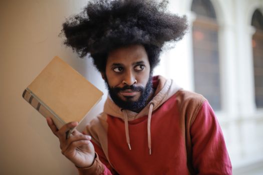 Young bearded ethnic male student with creative Afro hairstyle wearing hoodie sweatshirt looking away with doubt expression while taking old book against blurred exterior of aged university