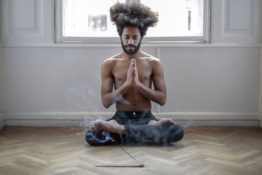 Shirtless man meditating in lotus pose indoors with incense smoke creating a serene atmosphere.