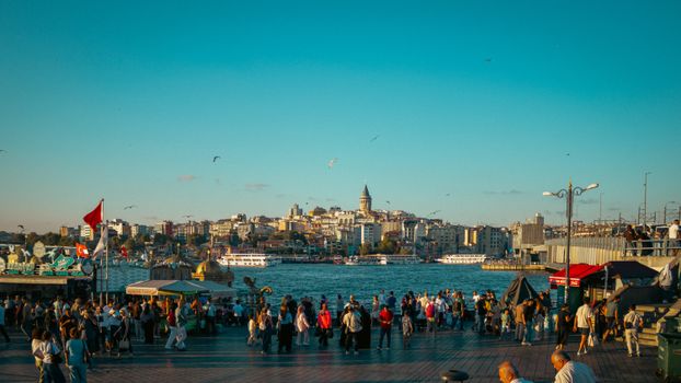 Crowded waterfront with Galata Tower in Istanbul under a clear blue sky.