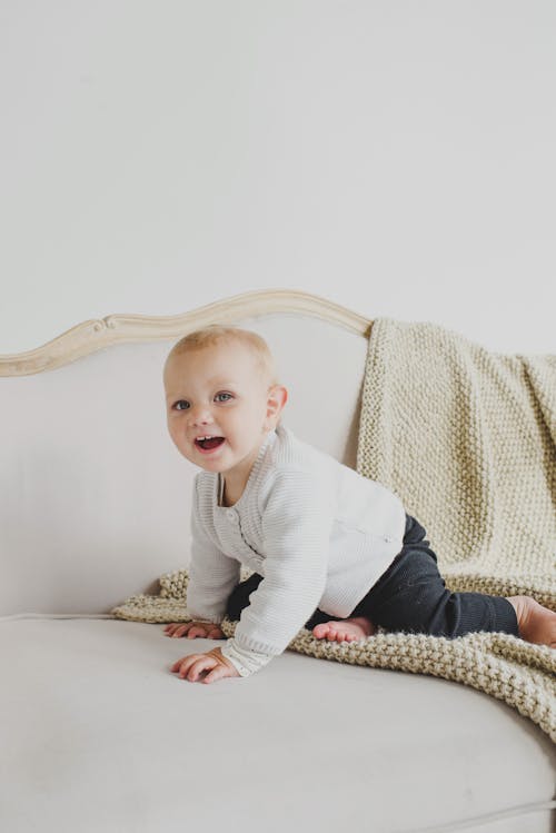 Happy little girl in casual clothes sitting barefoot on beige fluffy plaid placed on comfortable couch of classic style in light room with white wall and looking at camera in daylight