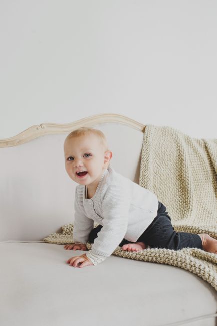 Happy little girl in casual clothes sitting barefoot on beige fluffy plaid placed on comfortable couch of classic style in light room with white wall and looking at camera in daylight