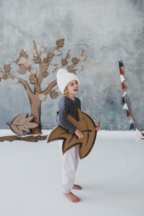 Free Full body delighted child in warm clothes and hat standing barefoot inside scenery room with artificial autumn tree and fallen leafs together with garland of multicolored flags with big brown leaf in hand and smiling looking away Stock Photo