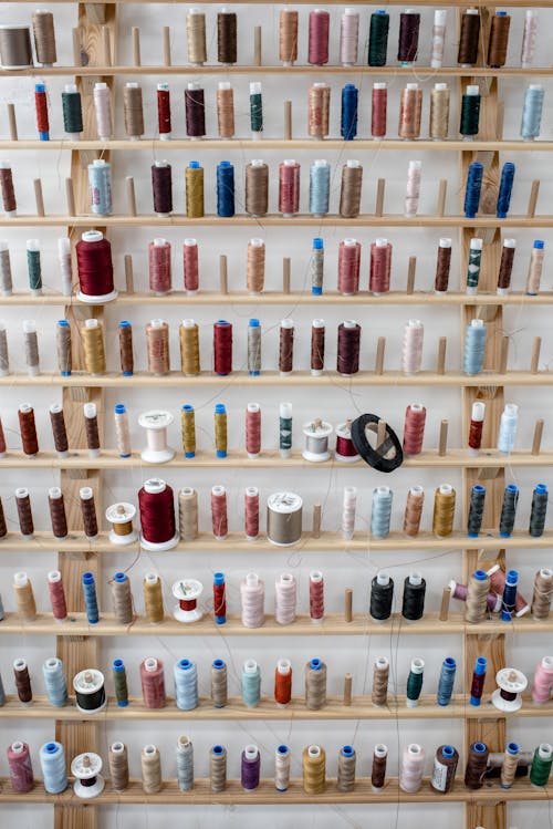 Stand with assorted colorful spools of thread for sewing placed near white wall inside dressmaker workshop or tailor atelier for fashion design and apparel creation