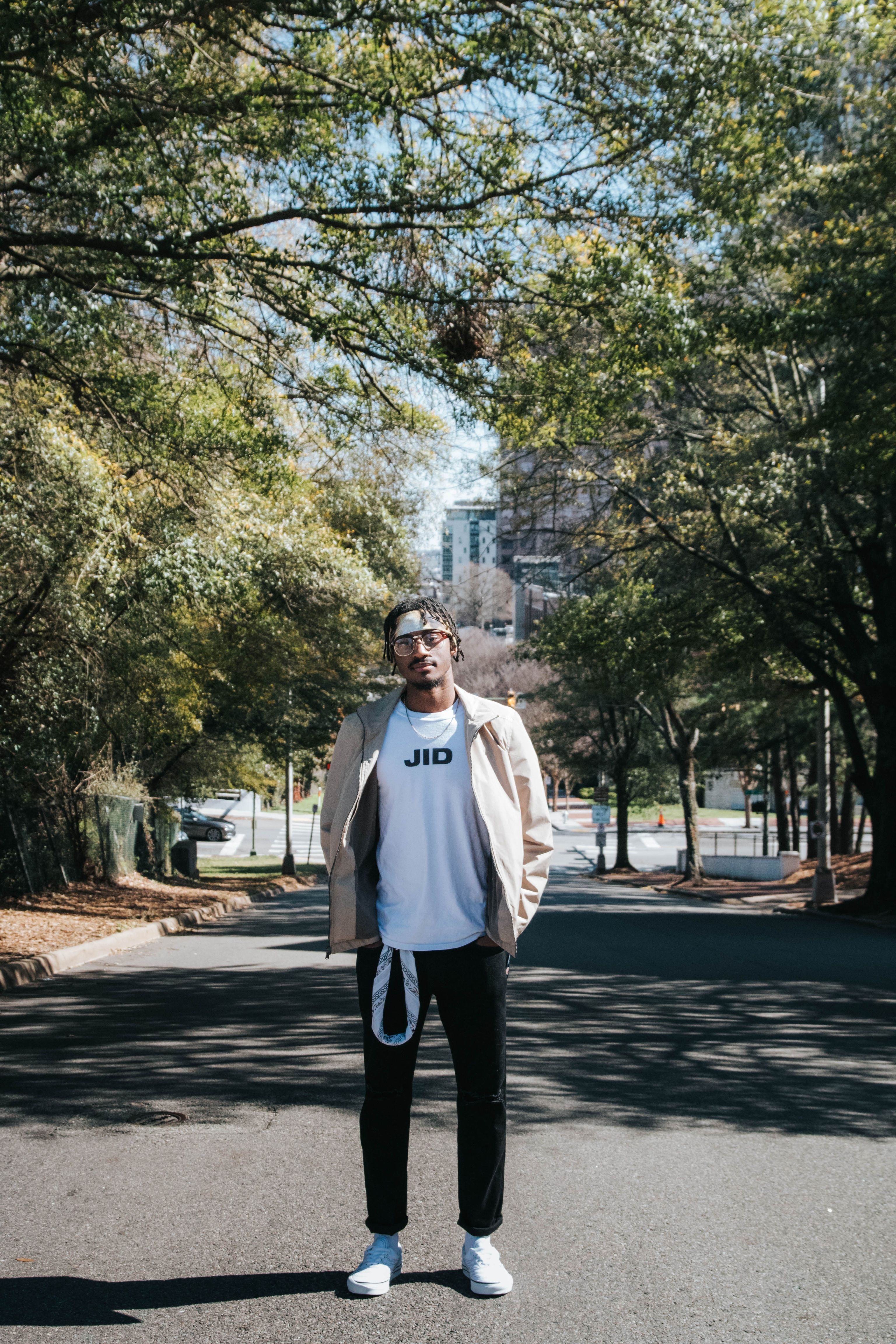 Young man wearing casual attire standing in a tree-lined street on a sunny day.