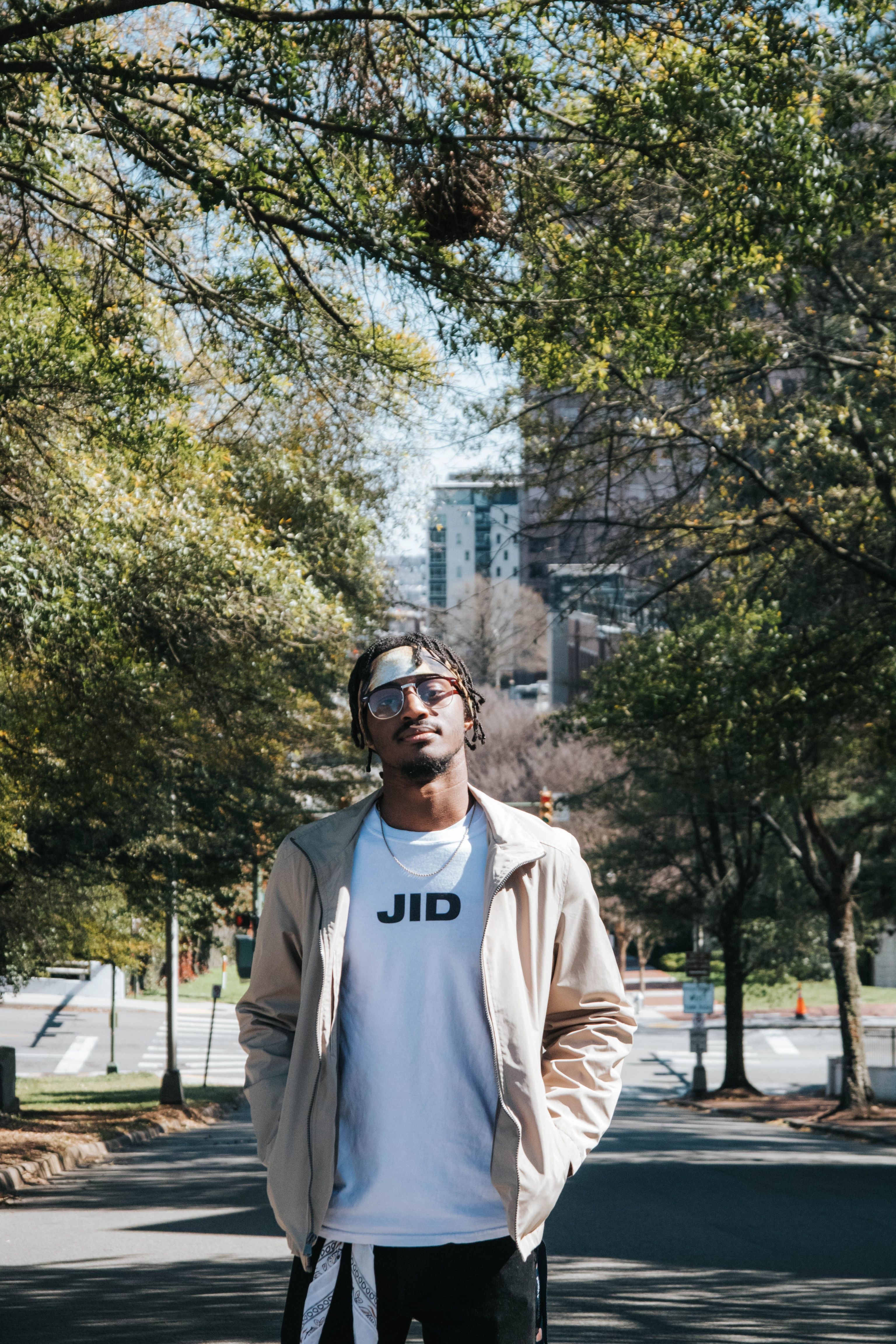 A fashionable young man with sunglasses poses confidently outdoors in a sunny urban park with city buildings in the background.