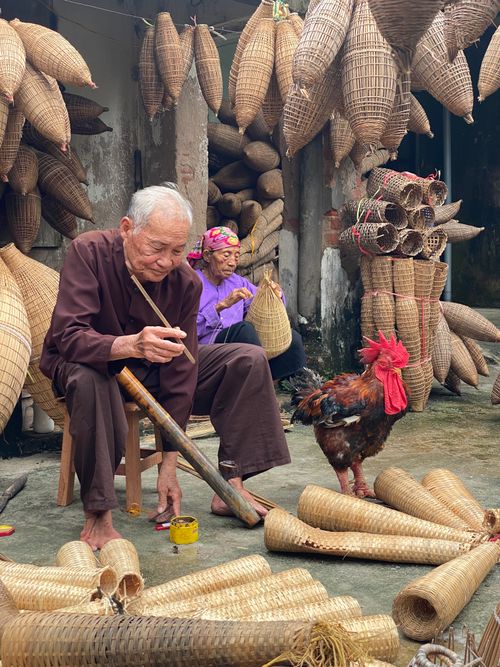 Free Elderly man skillfully weaving bamboo baskets surrounded by traditional crafts and a rooster. Stock Photo