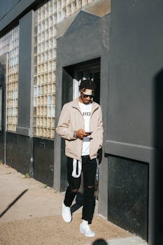 Man walks along city sidewalk, texting on phone, showcasing urban fashion and lifestyle.