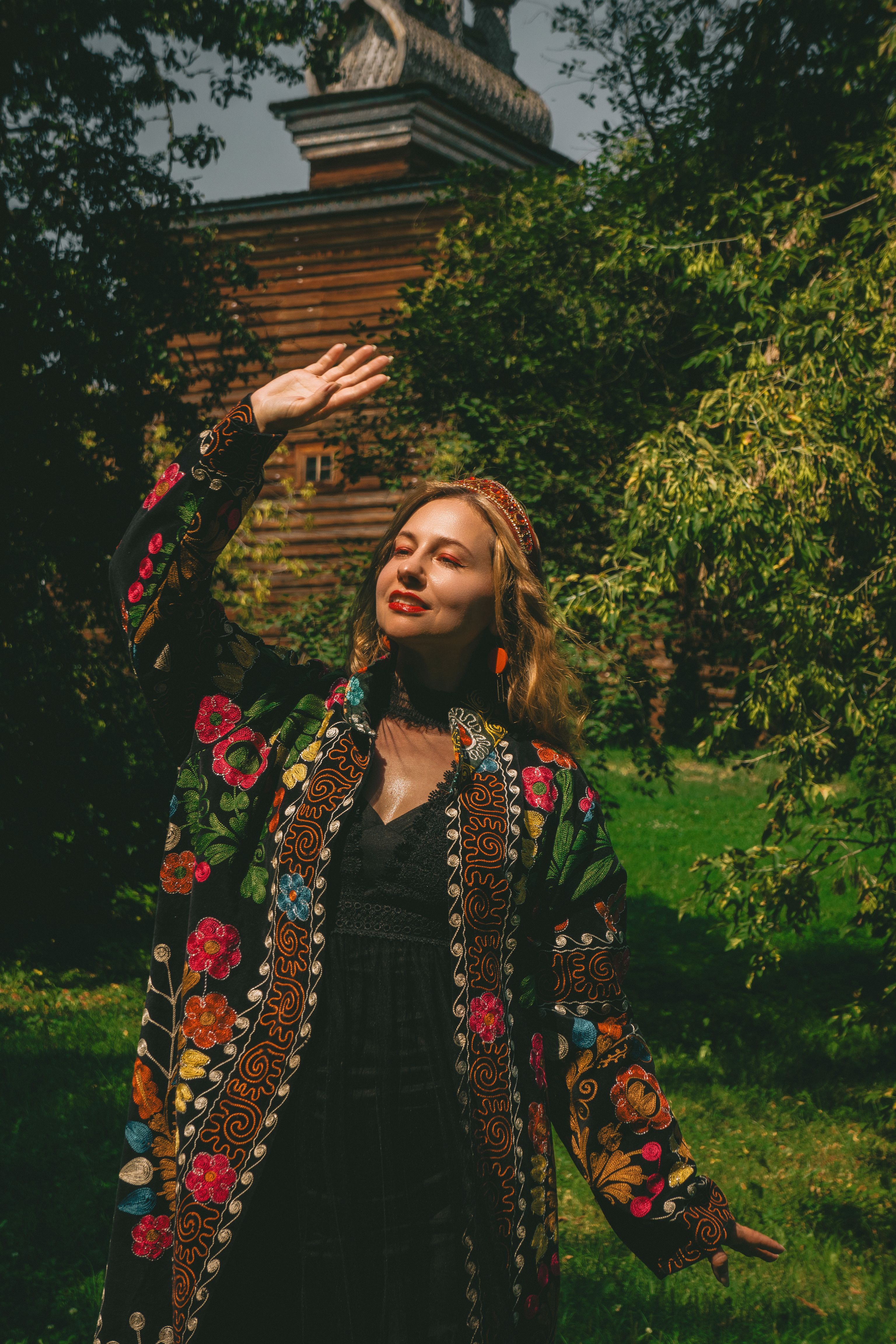 A woman in vibrant traditional floral attire enjoys a sunny day outdoors near a wooden structure.