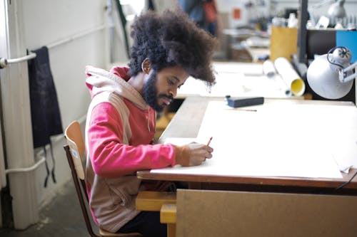 Man in Pink Jacket Sitting on Brown Chair Drawing on White Paper