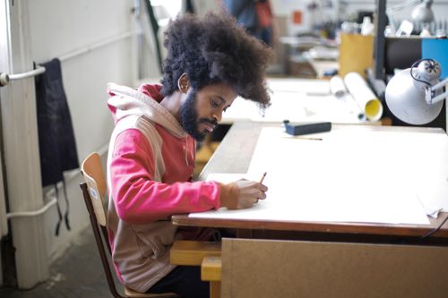 Free A dedicated artist with afro hair sketching at a desk in an inspiring indoor workspace. Stock Photo