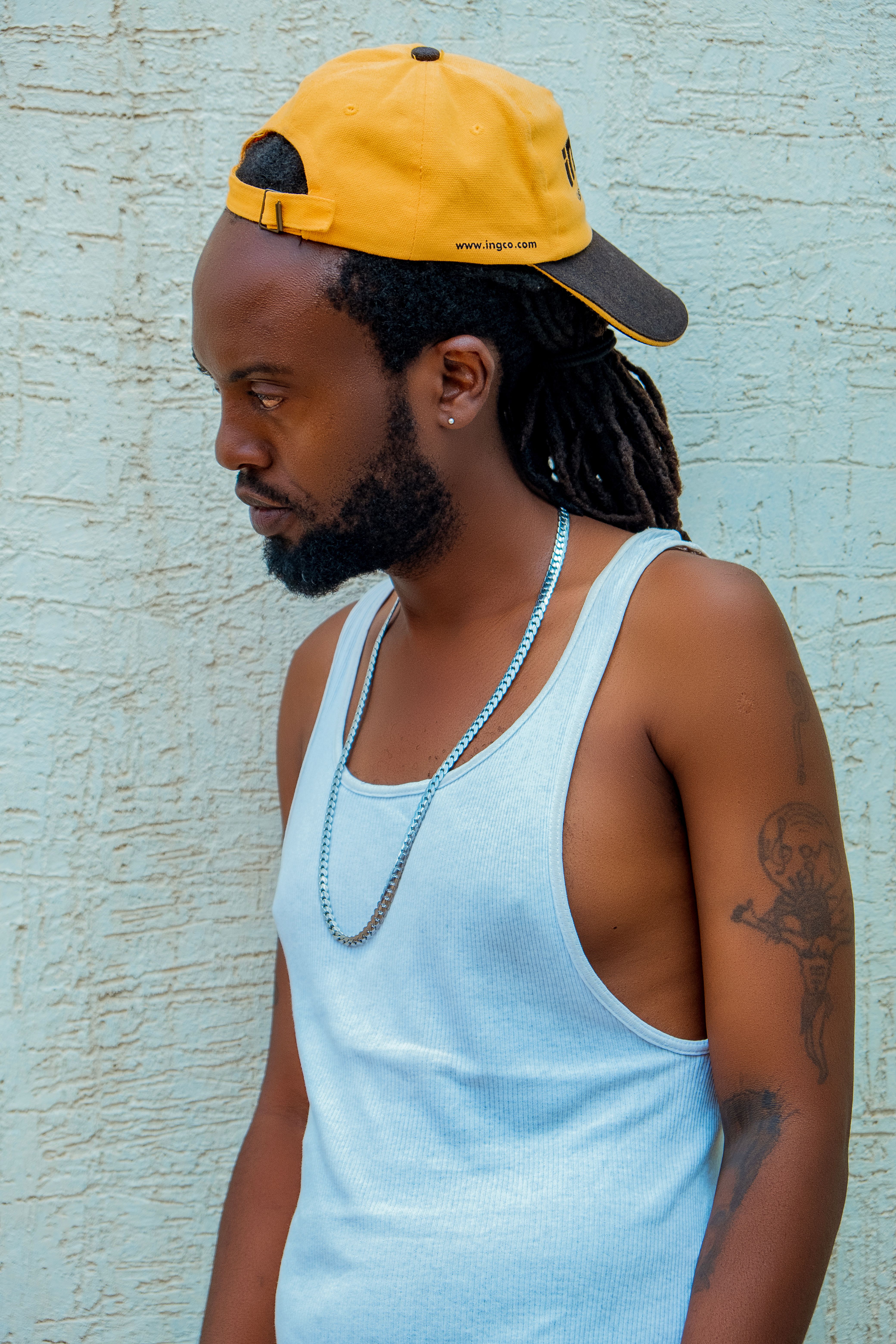 Man in a tank top and cap posing against a textured wall.